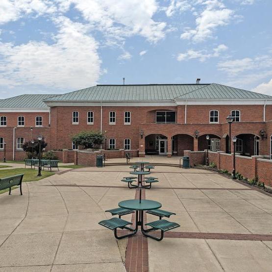 A wide two-story brick building with a pale green tiled roof. There is a courtyard with green tables to sit and eat at.